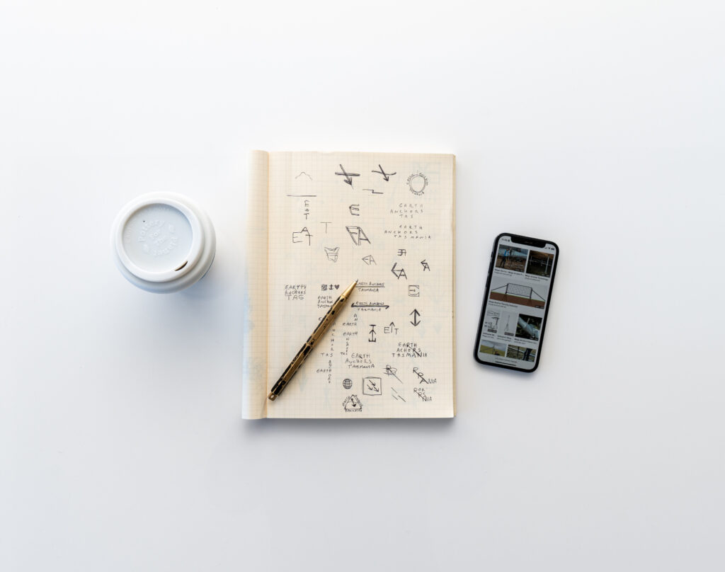 A notebook showing sketch ideation of Earth Anchors Tasmania's logo design. A coffee mug and iphone are next to the notebook